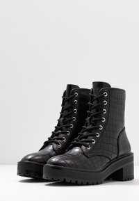 Zwarte veterlaarzen met een gestructureerd krokodillenpatroon, een dikke plateauzool en zilverkleurige oogjes. Gemaakt van synthetisch materiaal.