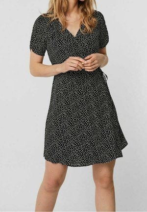 Femme portant une robe portefeuille noire à pois blancs, manches courtes et col en V, debout les mains jointes devant.