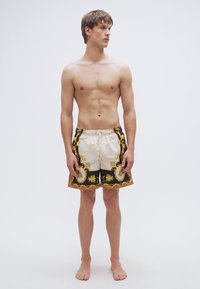 Κοντά μαγιό σε εκρού και μαύρο με περίπλοκα χρυσά σχέδια και κορδόνι στη μέση. Διαθέτουν άνετη εφαρμογή και μήκος μέχρι το γόνατο.