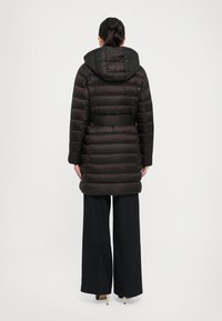 Kvinne iført svart belted pufferjakke med hette, vide svarte bukser og beige høyhælte sko, vender bort fra en enkel bakgrunn.