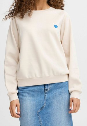 Femme portant un sweat crème avec un petit cœur bleu sur la poitrine gauche et une jupe en denim bleu clair avec une couture asymétrique.