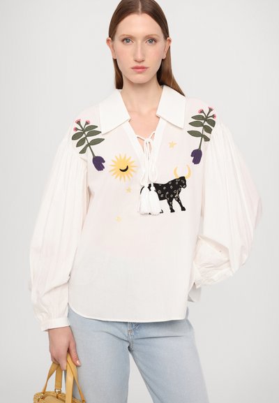 Weiße Bluse mit großem Kragen, verziert mit bunten gestickten Motiven, darunter Blumen, eine Sonne und ein schwarzer Stier. Fließende Ärmel und Fransen Details.