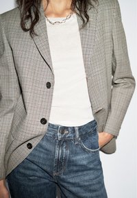 Kvinna iklädd en rutad blazer, vit t-shirt, blå jeans och en silver kedja, med en hand i fickan mot en vit bakgrund.