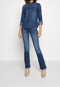 Camicia di jeans con maniche lunghe, chiusura con bottoni, due tasche sul petto; abbinata a jeans blu svasati e stivaletti beige con tacco a blocco.