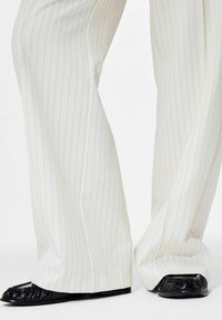 Pantalon large blanc à rayures fines, associé à des chaussures plates noires texturées sur fond blanc.