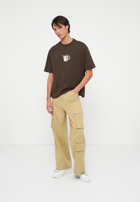 Καφέ βαμβακερό μπλουζάκι με γραφικό καφέ, συνδυασμένο με μπεζ cargo παντελόνι με πολλές τσέπες και χαλαρή εφαρμογή. Λευκά αθλητικά παπούτσια.