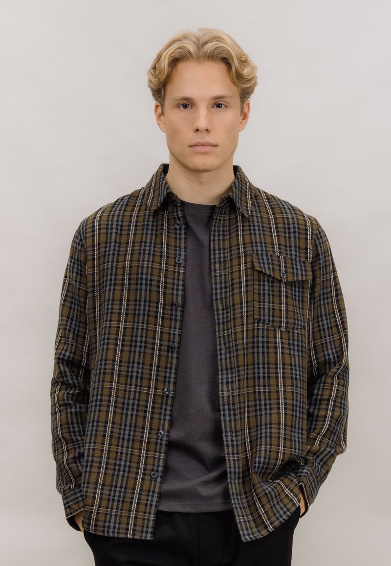 Brun og svart rutete flanellskjorte med knappert front, brystlomme og lange ermer over en grå crew neck t-skjorte.