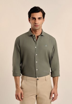 Camisa de manga larga verde oliva claro, con botones, patrón texturizado, bolsillo en el pecho y puños enrollados. Presenta botones blancos y un pequeño logo.