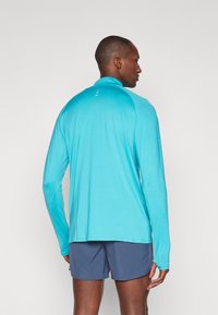 Uomo che indossa una maglietta sportiva a maniche lunghe turchese e pantaloni corti blu navy, in piedi con la schiena rivolta verso uno sfondo chiaro e semplice.