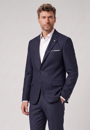 Man with short curly hair wearing a navy blue suit and white shirt, standing with one hand in pocket against a plain background.