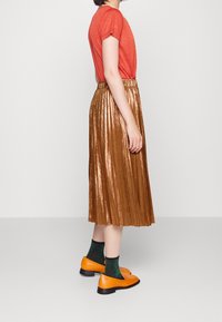 En rostorange t-shirt, en metallisk veckad bronskjol, gröna glittriga strumpor och orangea platta skor, sedd från sidan mot en enkel bakgrund.