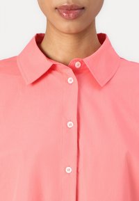 Chemise boutonnée corail avec un col structuré, ornée de boutons roses et d'une texture de tissu lisse et léger.