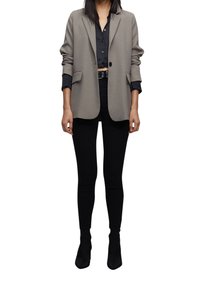 Blazer gris clair avec épaules structurées, fermeture à un bouton et poches, porté sur une chemise noire boutonnée et un pantalon noir.