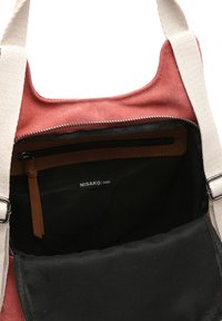 Röd tygväska med insidan i svart, brun läderdetalj och dragkedja. Etiketten säger "MISAKO." Justerbar krämfärgad rem synlig.