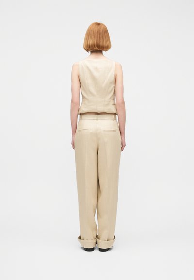 Άτομο με κοντά κόκκινα μαλλιά, που φοράει μπεζ αμάνικο τοπ και παντελόνες με φαρδιά μπατζάκια, στέκεται πλάτη σε απλό λευκό φόντο.