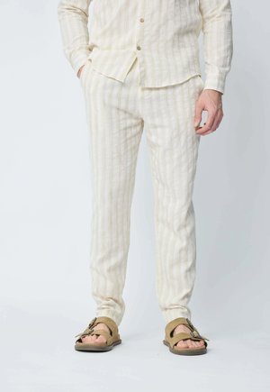 Ensemble de chemise et pantalon rayé beige clair, fabriqué en tissu texturé. Pantalon slim, porté avec des sandales brunes avec une boucle.