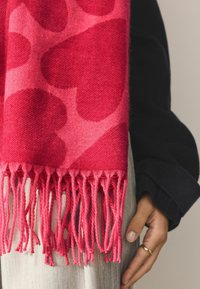 Bufanda roja con un patrón de corazones, hecha de tela suave. Presenta bordes con flecos y un diseño degradado, mostrada sobre un fondo neutro.