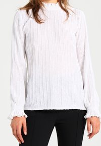 Blusa bianca e tessurizzata con design rigato verticale, maniche lunghe, polsini volant e collo alto. Indossata con pantaloni neri.