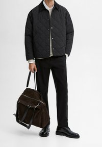 Musta tikattu takki, jossa kaulus, harmaa neule alla, mustat housut ja mustat kengät; kädessä ruskea mokkanahkainen laukku hopeisilla metalliosilla.