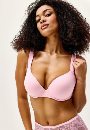 Nainen, jolla on kiharat mustat hiukset, pukeutunut vaaleanpunaiseen toppattuun rintaliiviin ja siihen sopiviin pitsialushousuihin yksiväristä taustaa vasten.