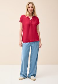 Rotes Kurzarmoberteil mit V-Ausschnitt und Knopfdetails, kombiniert mit hellblauen weit geschnittenen Jeans und beigen Sneakers vor einem neutralen Hintergrund.
