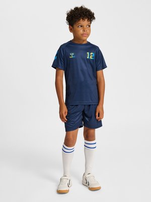 Junge trägt ein marineblaues Sporttrikot mit der Nummer 12, passende Shorts, weiße knielange Socken mit blauen Streifen und weiße Turnschuhe, steht und schaut nach rechts.