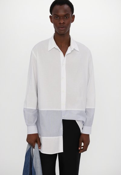 Chemise blanche à boutons avec des rayures verticales et une section inférieure à rayures bleues. Elle possède un col classique et des manches longues. Coupe légèrement oversize.