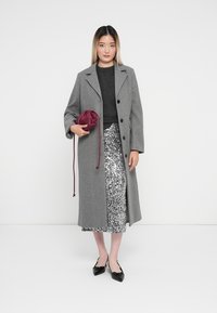 Grijze wollen jas met grote knopen, gedragen over een donkere trui, gecombineerd met een glanzende zilveren paillettenrok en een kastanjebruine clutch. Zwarte ballerina's.