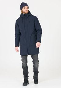 Parka impermeable navy con capucha ajustable, botones a presión y bolsillos laterales, combinada con jeans oscuros y botas de senderismo negras.