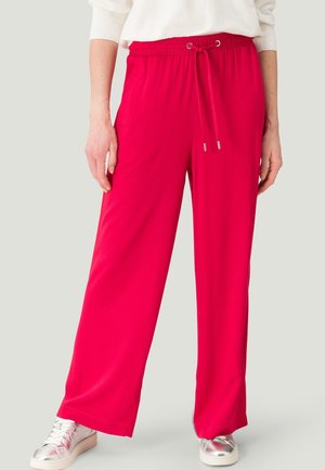 Dolna część ciała osoby noszącej jaskrawo czerwone szerokie spodnie z wiązanym pasem, biały sweter i srebrne sneakersy na neutralnym tle.