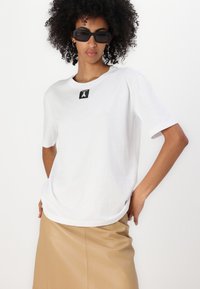 Balta kokvilnas T-kreklā ar īsām piedurknēm, apaļo kakla izgriezumu un melnu kvadrātu birku uz krūtīm, apvienota ar bēša svārkiem.