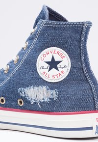 Vysoké džínové tenisky Converse s opotřebeným záplatou, bílou logovou nášivkou, stříbrnými kovovými očky a červeným akcentem na podrážce.