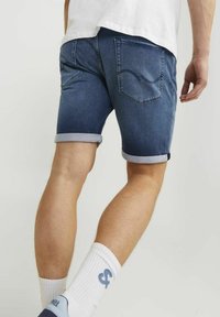Man som bär uppvikta mörkblå denimshorts, vita strumpor med blå logotyp och marinblå sneakers, stående mot en enkel ljus bakgrund.