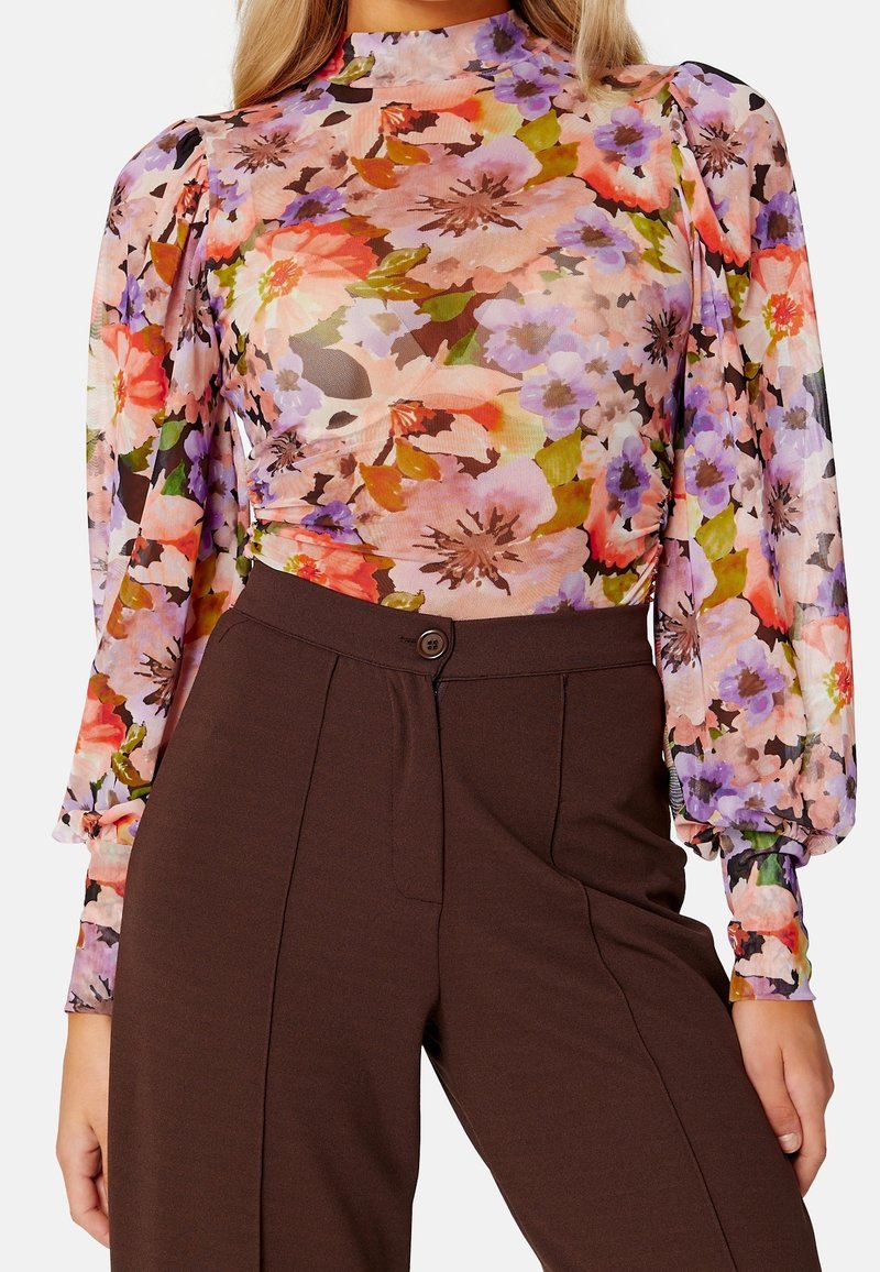 Haut à manches longues floral avec un col haut ; présente des fleurs roses, violettes et oranges vives sur un tissu transparent ; associé à un pantalon taille haute marron.