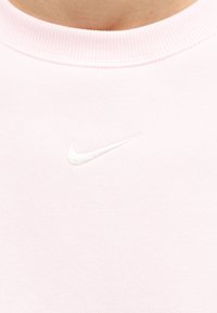 Κοντινό πλάνο ενός ανοιχτό ροζ φούτερ με στρογγυλή λαιμόκοψη και λευκό λογότυπο Nike swoosh κεντημένο στο στήθος.