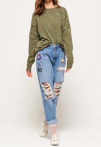 Sudadera de manga larga color verde oliva con diseño de salpicaduras, combinada con jeans rotos de color azul claro que tienen parches coloridos y puños enrollados.
