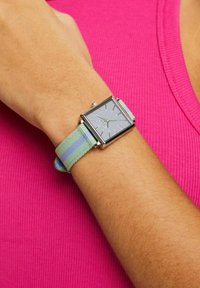Firkantet sølvur med en grå urskive og sølvfarvede timemarkører, monteret på et strap af stribede stof i grøn og lavendel.