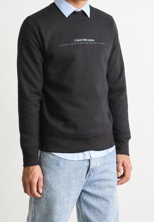 Mężczyzna ubrany w czarną bluzę Calvin Klein na niebieską koszulę z kołnierzykiem, w połączeniu z jasnoniebieskimi jeansami, stoi na neutralnym tle.