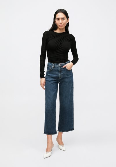 Črna majica z dolgimi rokavi iz teksturirane tkanine, kombinirana z modrimi kavbojkami z visokim pasom in širokimi hlačnicami ter belimi petami z ostrim prstom.