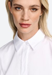 Biała koszula zapinana na guziki z klasycznym kołnierzykiem, gładką teksturą i starannymi ściegami. Modelka ma jasne blond włosy i naturalny makijaż.
