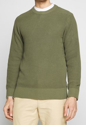 Man in een olijfgroen gebreide trui en beige broek, staand met ontspannen armen tegen een effen lichte achtergrond.