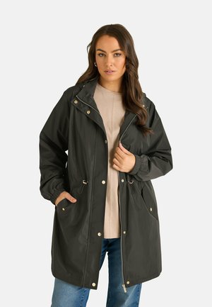 Femme aux longs cheveux bruns portant un imperméable noir à capuche par-dessus un haut beige et un jean bleu, debout avec une main dans la poche et l'autre ajustant son manteau.