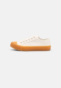 Canvas sneaker in cream with a ribbed orange rubber sole and toe. Features five eyelets and minimalistic design without prominent details.