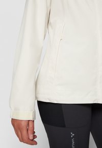 Veste légère de couleur crème avec une coupe ajustée, une texture douce, des poignets réglables à velcro et un logo sur l'ourlet. Adaptée pour une tenue active.