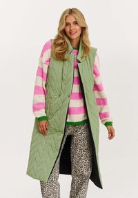 Gepolsterte, ärmellose lange Weste in Hellgrün mit schwarzem Futter. Über einem pink-weiß gestreiften Pullover und gemusterten Hosen getragen.