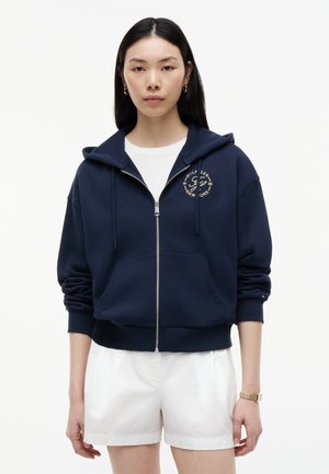Jonge vrouw draagt een marineblauwe zip-up hoodie met logo en witte korte broek, staand tegen een effen witte achtergrond.