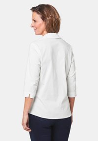 Blusa bianca in cotone con maniche a tre quarti, colletto e spacchi laterali sui polsini. Sul retro presenta un design minimale e liscio.