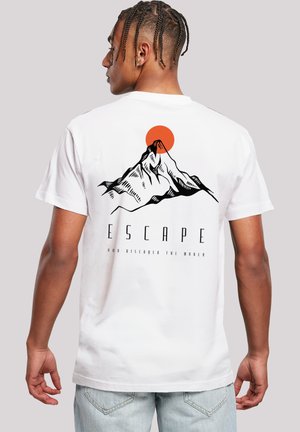 Junger Mann mit geflochtenem Haar, der ein weißes T-Shirt trägt, das ein schwarzes Bergmotiv und eine orange Sonne zeigt. Auf der Rückseite steht das Wort "ESCAPE".