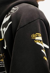 Felpa con cappuccio nera con stampe grafiche bianche e gialle, dettagli testuali e una texture morbida. Il design include vari elementi astratti.