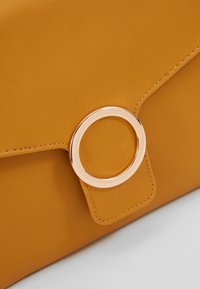 Żółta skórzana kopertówka o gładkiej teksturze, z wyraźnym okrągłym detalem w kolorze złotym oraz przeszyciami jako akcentami.